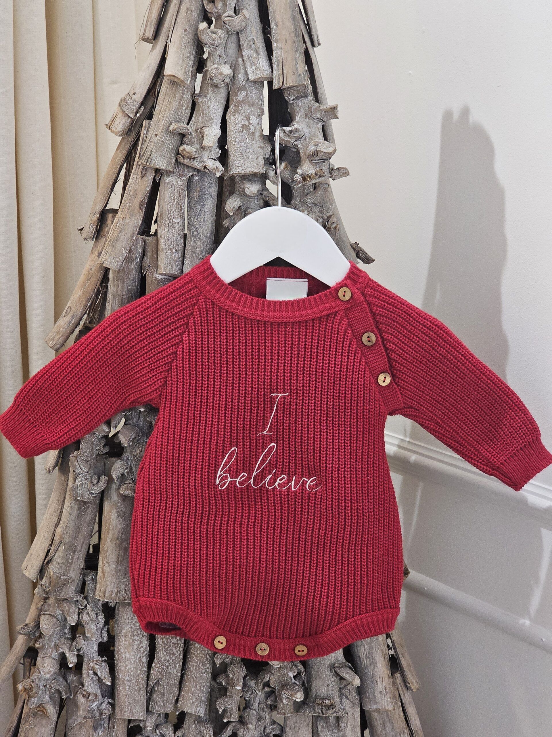 the 'I Believe' Red Christmas Romper displayed hanging from a decorative stick tree
